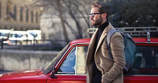 30-something man wearing sport coat and carrying a backpack standing next to car