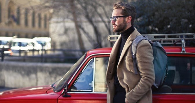 30-something man wearing sport coat and carrying a backpack standing next to car